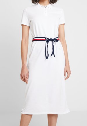 Frau trägt ein weißes Polokleid mit kurzen Ärmeln und einem marineblauen, roten und weißen gestreiften Gürtel, der in einer Schleife an der Taille gebunden ist.