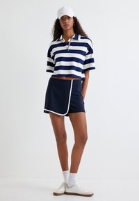 Polo court rayé bleu marine et blanc avec un col et une fermeture éclair ; associé à une jupe portefeuille bleu marine avec des passepoils blancs et des baskets sportives.