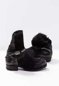 Par sorte ruskindsankelstøvler med foret pels og teksten "BELIEVE IN YOU" på en rem omkring anklen, vist mod en enkel hvid baggrund.