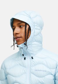 Polstret, lyseblå jakke med høj krave og fastgjort hætte. Har en quiltet tekstur og sorte justerbare snore ved lynlåsen.