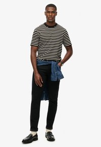 Homme portant un t-shirt rayé noir et blanc, un pantalon noir, des mocassins noirs avec des chaussettes blanches, et une veste en denim bleue attachée autour de la taille.