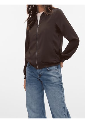 Vrouw draagt een zwarte bomberjack met rits over een wit shirt en lichtblauwe, high-waisted rechte jeans, hand in de zak.