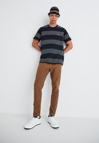 Randig navy- och vit kortärmad t-shirt med rund halsringning, parat med tan-färgade byxor och vita sneakers. Accentuerad med en randig bucket-hatt.