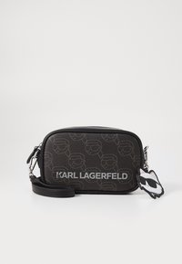 Črna teksturirana torba preko telesa z embosiranim vzorcem obrazov, srebrnim logotipom in obeskom v obliki mačke, pritrjenim na trak.