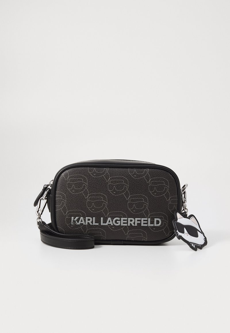 Črna teksturirana torba preko telesa z embosiranim vzorcem obrazov, srebrnim logotipom in obeskom v obliki mačke, pritrjenim na trak.