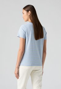 Striped T-shirt in light blue with white stripes, short sleeves, round neckline; fabric appears soft and casual. Worn with cream-colored pants.
