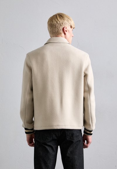 Μπεζ σακάκι από μείγμα μαλλιού με ριγωτούς καρπούς, διαθέτοντας κλασικό γιακά και διακριτή υφή, συνδυασμένο με σκούρα τζιν σε ουδέτερο φόντο.