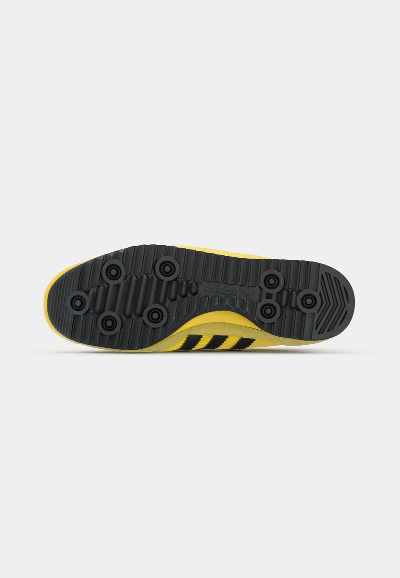 Suela de zapato Adidas con una combinación de colores amarillo y negro, que presenta una superficie de goma negra texturizada y tres rayas negras.