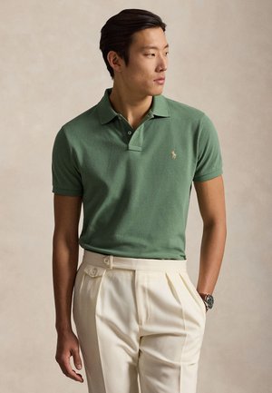 Homme portant un polo vert à manches courtes et un pantalon crème taille haute, regardant sur le côté avec une main dans la poche.