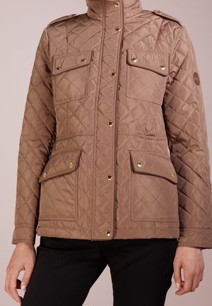 Veste matelassée marron avec quatre poches à rabat et boutons-pression dorés, portée sur un pantalon noir.