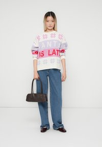 Strickpullover mit weißen, lavendelfarbenen und pinken Schneeflockenmuster sowie roten Text; kombiniert mit blauen Jeans und einer dunkelbraunen Handtasche.