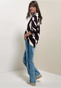 Blusa a maniche lunghe con motivo zig zag in viola scuro e crema, abbinata a jeans chiari a zampa d'elefante e sneakers beige. Tessuto morbido.