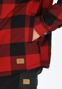Rood en zwart geruit overhemd met een knoopmanchet, zijzak en merkpatches op de zoom en zak, gemaakt van dik stof.
