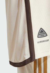 Lichtbeige sportshirt van textuurstof, met bruine accenten, een driehoekige logo patch en het "AEROREADY" label aan de zijkant.