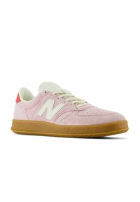 Baskets New Balance en daim rose clair avec lacets blancs, logo blanc, semelle en gomme et accent rouge sur le talon, présentées avec un léger angle de côté.