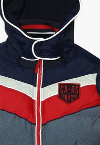 Vadderad jacka med en marinblå, röd och grå färgschema. Har en dragkedja fram, huva, randiga detaljer och en broderad logotyp. 