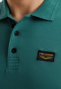Tyrkysové polo triko vyrobené z texturované látky, s černým límečkem a třemi knoflíky. Výrazná černá nášivka značky se zlatým textem.