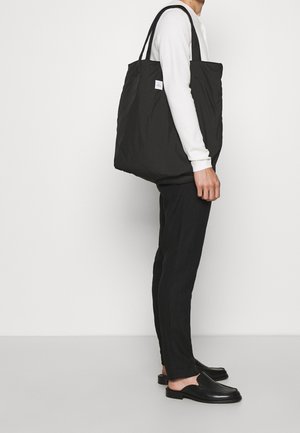 Mann trägt schwarze Hose, weißes Langarmshirt, schwarze Slipper und trägt eine große schwarze Tragetasche über der Schulter vor weißem Hintergrund.