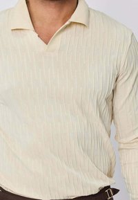 Polo à manches longues de couleur crème, présentant un motif texturé, un col en V et une coupe décontractée. Le tissu est lisse, léger et polyvalent.