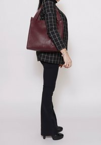 Bordeaux leren tote bag met textuur, driehoekige vorm en dubbele handgrepen, gepresenteerd naast een model in een zwart geruit top.