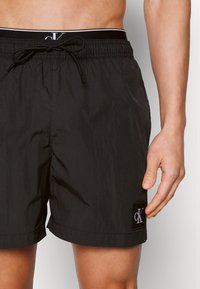 Shorts de bain noirs avec une taille à cordon de serrage, fabriqués en tissu léger et lisse, et ornés d'une petite patch logo CK en bas à gauche.
