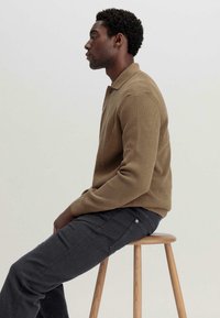 Brauner gerippter Strickpullover mit Kragen und Knopfdetails, kombiniert mit dunkelgrauen Hosen, auf einem Holzstuhl sitzend, seitlich betrachtet.