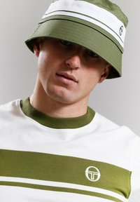 Cappello a secchiello verde con strisce orizzontali bianche, indossato sopra una t-shirt bianca con strisce verdi e un logo circolare sul petto.