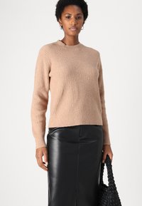 Beige gerippter Strickpullover mit rundem Halsausschnitt und langen Ärmeln, kombiniert mit einem schwarzen Ledrrock und einer schwarzen geflochtenen Tasche.