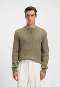 Olivgrüner Strickpullover mit Fischgrätenmuster, Rundhalsausschnitt und gerippten Bündchen. Über weißen Hosen getragen, zeigt er eine lässige Passform.