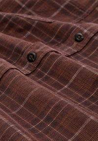 Chemise en plaid marron avec un tissu texturé. Équipée de boutons noirs et de fines lignes blanches dans le motif. Design structuré avec une surface lisse.