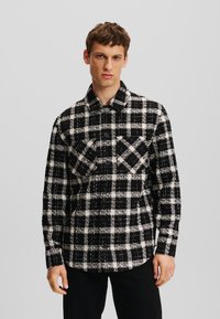 Camisa de franela a cuadros en blanco y negro con un diseño de botones, dos bolsillos en el pecho y un cuello. La textura de la tela parece suave y texturizada.