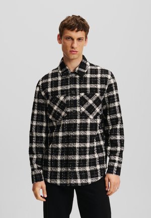 Camisa de franela a cuadros en blanco y negro con un diseño de botones, dos bolsillos en el pecho y un cuello. La textura de la tela parece suave y texturizada.