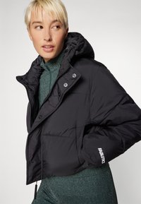 Veste courte noire en duvet avec capuche, présentant une texture brillante et un design matelassé. Comprend des fermetures à pression et un logo visible sur la manche.