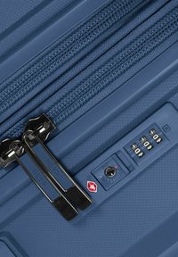 Valigia rigida blu con superficie testurizzata, doppie cerniere e lucchetto a combinazione approvato dalla TSA. Presenta dettagli metallici lucidi.