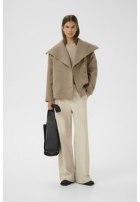 Beige wool jacket with wide collar, button closure, and over-sized fit, paired with cream wide-leg trousers and a black tote bag.