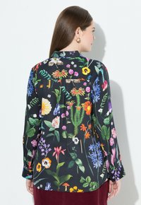 Una donna con lunghi capelli castani indossa una blusa nera con motivi floreali colorati, piante, dadi e chiavi, abbinata a una gonna rosso scuro.
