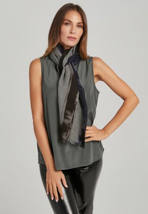 Frau trägt ein ärmelloses graues Oberteil, schwarze glänzende Hose und einen schwarz-weißen gemusterten Schal, posiert mit einer Hand in der Hüfte.