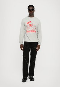 Gray sweatshirt with red graphic text and illustration, paired with black jeans featuring gold studded accents. Model wears black sunglasses and shoes.