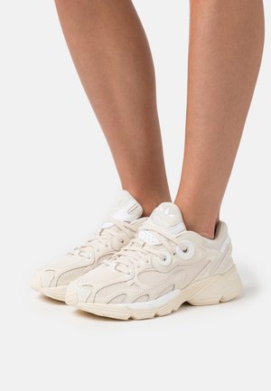 Baskets Adidas beige clair avec détails en mesh et cuir portées aux pieds sur fond blanc.