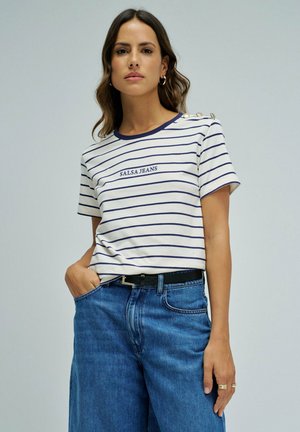 Mujer con camiseta de rayas blancas y azul marino "SALSA JEANS" con botones en el hombro, pantalones vaqueros azules, cinturón negro y pendientes de aro dorados.