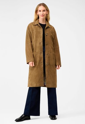 Vrouw draagt een lange bruine jas met knopen, een zwart shirt, donkere wijde broekspijpen en zwarte loafers, staand tegen een witte achtergrond.