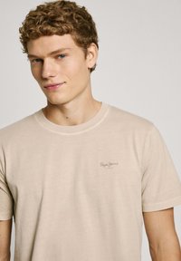 Camiseta beige de manga corta con cuello redondo y un logo sutil en el pecho. Tejido suave con una textura lisa.