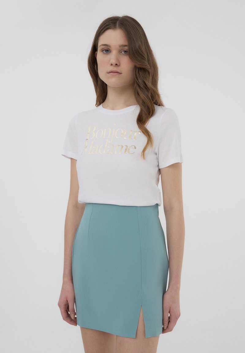 T-shirt bianco con testo dorato "Bonjour Madame", abbinato a una gonna corta e aderente in teal con uno spacco laterale. Tessuto liscio, design contemporaneo.