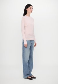 Light pink long-sleeve top with a fitted body, paired with high-waisted wide-leg blue jeans. Model stands sideways against a white backdrop.