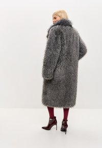 Grå pelsfrakke i imiteret pels med et voluminøst design, høj krave og lange ærmer. Båret over bordeauxfarvede strømpebukser og blanke ankelstøvler med hæl.