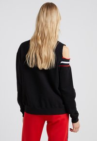Sweatshirt noir avec un décolleté tombant sur l'épaule. Présente des accents rayés rouge, blanc et noir sur les manches. Tissu doux et coupe décontractée.