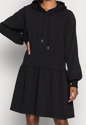 Kobieta w czarnej, długiej sukience z kapturem, z długim rękawem i sznureczkami do regulacji, o luźnym kroju, z marszczoną talią i mankietami na rękawach.
