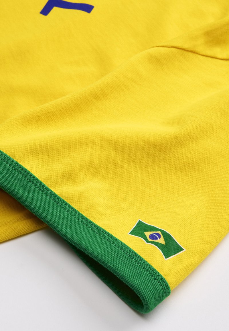 Tissu jaune avec une bordure verte présentant un petit emblème du drapeau brésilien près du bord, probablement une partie d'un vêtement.