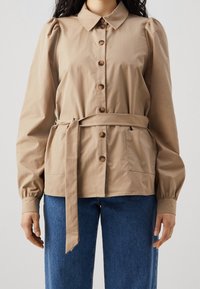 Ljust brunt skjorta med puffärmar och knytbälte, bärs över blå jeans. Skjortan har en krage och en bröstficka.