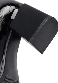 Zapato de charol negro con un tacón grueso negro. Textura suave con un acabado brillante y sutiles detalles de costura cerca de la suela.
